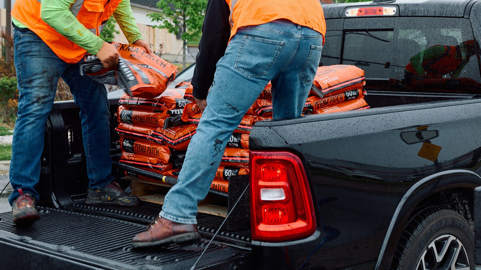 Two road crew members in high-visibility vests unloading 60 lb bags of Perma-Patch from the bed of a pickup on a wet street