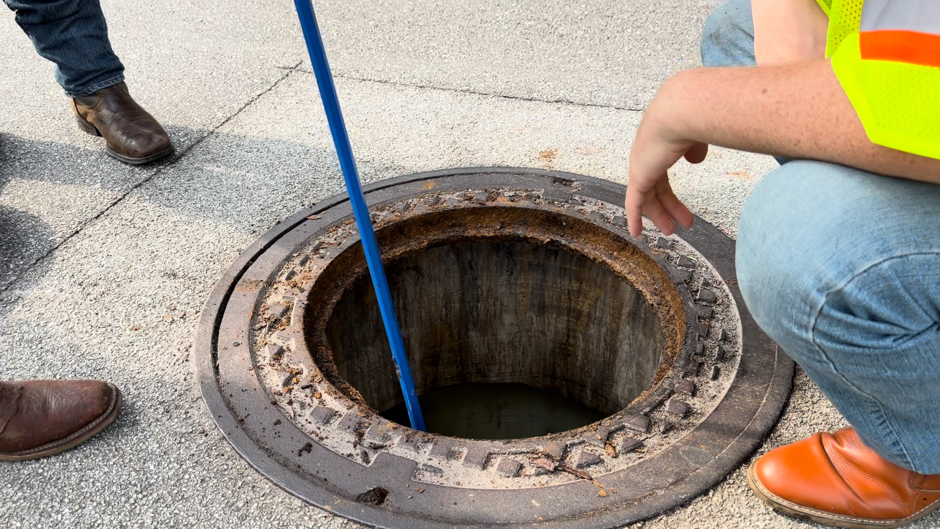 Crew member pointing into an open manhole frame while a probe is lowered down the chimney